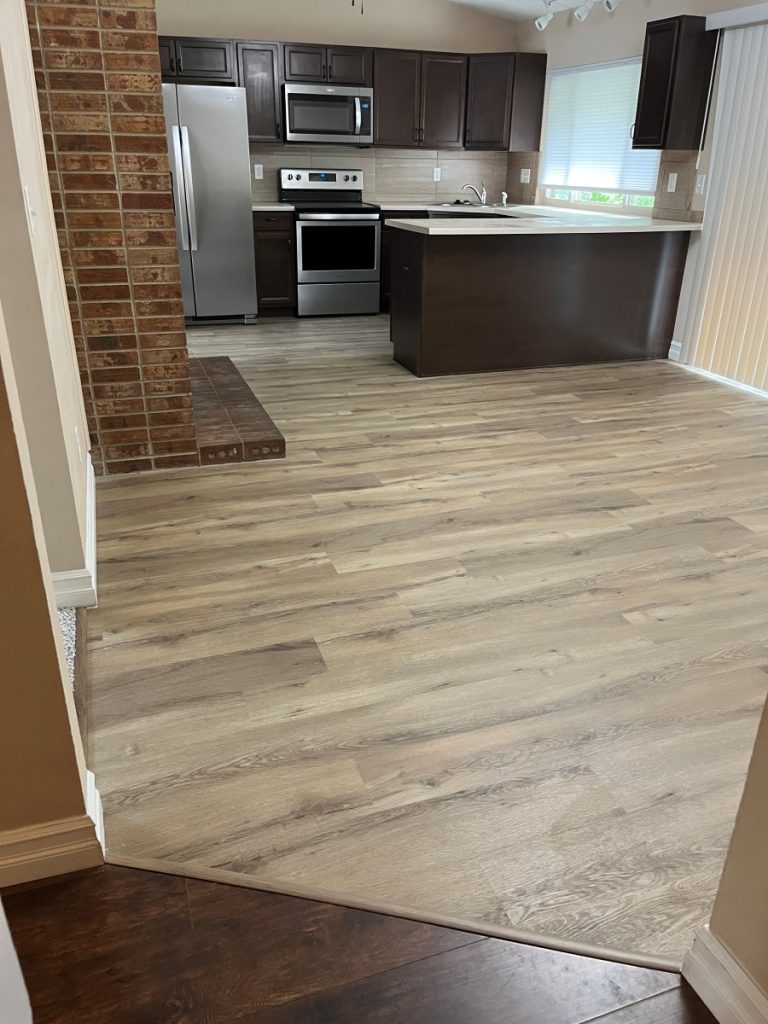 Modern kitchen with stainless steel appliances, dark wood cabinets, a light wood floor, brick accent wall, and an island with a light countertop. Natural light comes in through a window with blinds.