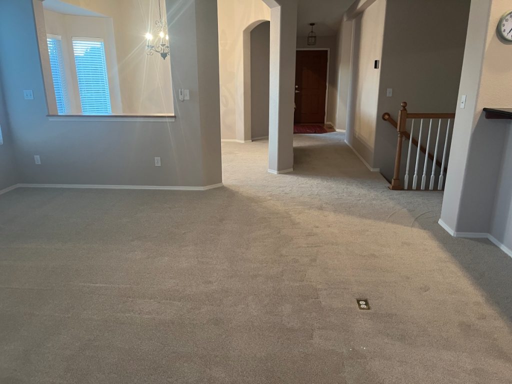 A spacious, empty room with light beige carpet, neutral walls, a staircase with a wooden handrail, a window, and a hanging chandelier. The room leads to a hallway and a front door in the background.