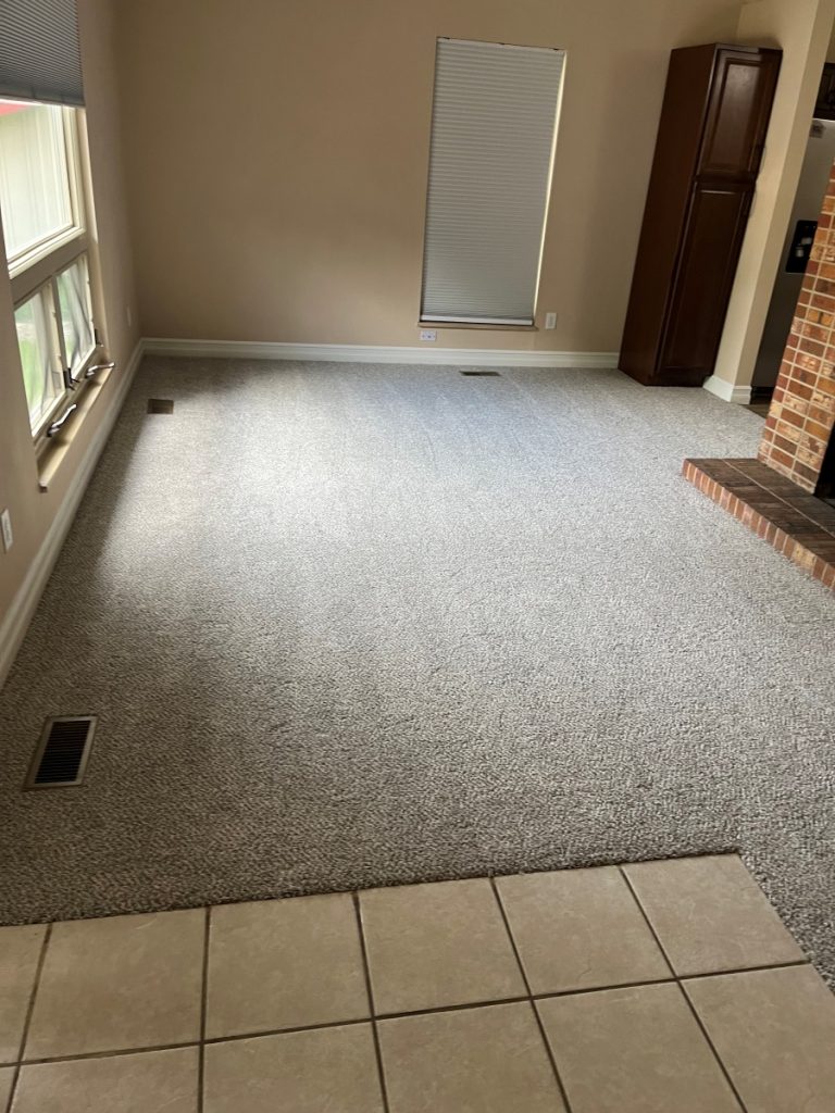 A carpeted living room with beige walls, windows on the left, a closed window blind, a brown cabinet in the corner, and a brick fireplace on the right; tile flooring is visible in the foreground.