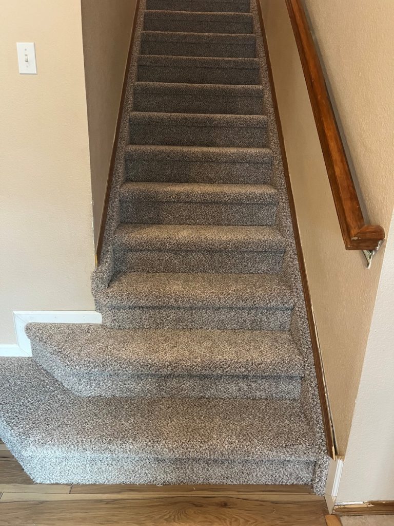 A carpeted staircase with beige walls, a wooden handrail on the right, and a small landing at the base where the bottom steps curve outward. The floor at the bottom of the stairs is light wood.