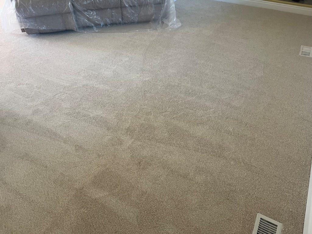 Beige carpet with visible vacuum lines and a subtle trail, with a couch partially wrapped in plastic in the background and a floor vent in the foreground.