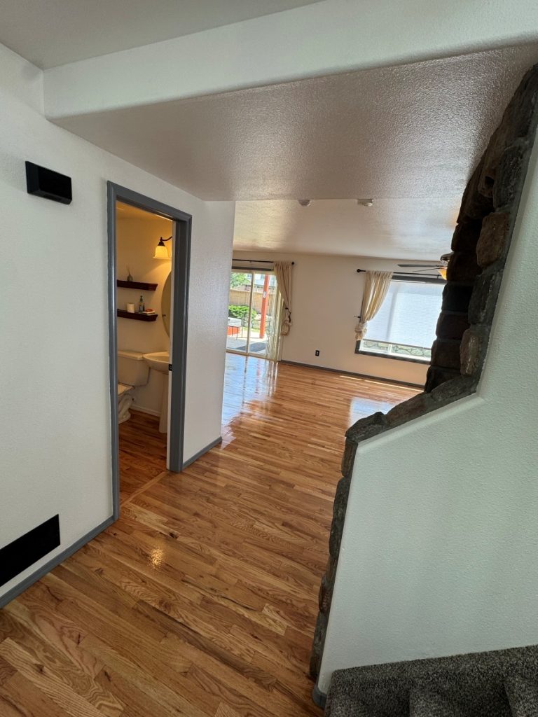 A hallway with polished wooden floors leads to a bright living area with large windows and curtains. To the left is a small bathroom with shelves, and a stone-accented wall is visible on the right near carpeted stairs.