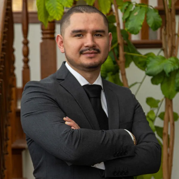 Jorge in a dark suit and black tie stands with arms crossed, smiling confidently. He is indoors, near a wooden staircase and large green leafy plants.