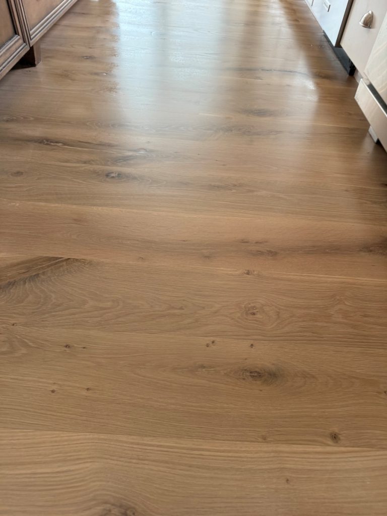 Light brown wooden floor with a smooth finish and visible natural grain patterns, knots, and textures, located along a hallway between cabinets and appliances.