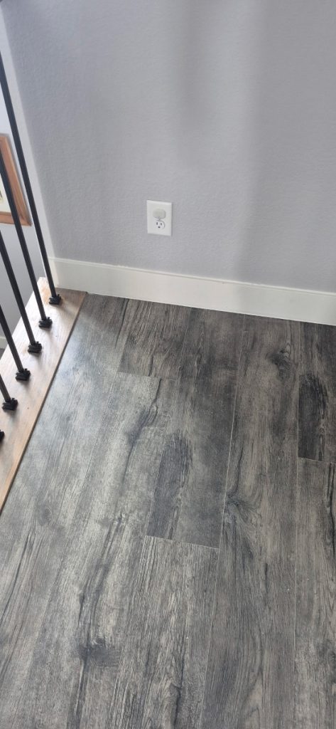 A section of a room with gray wood-patterned flooring, a white electrical outlet on a light gray wall, white baseboards, and a black metal railing on the left next to a wooden stair edge.