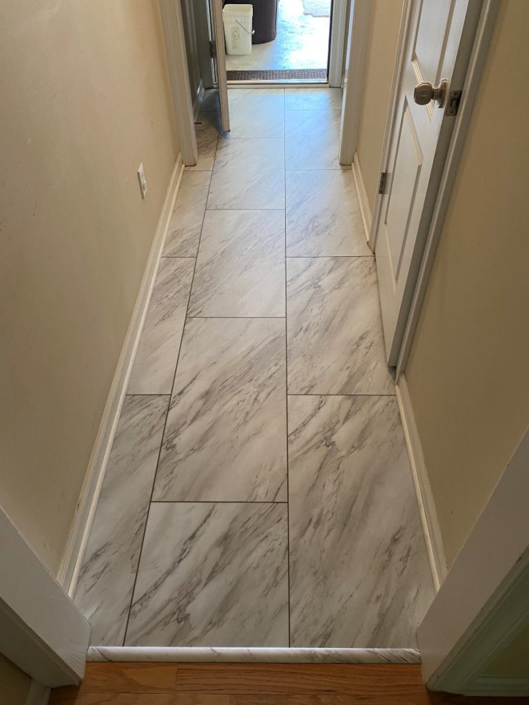 A narrow hallway with light-colored, marble-look floor tiles, cream walls, white trim, and a closed door on the right. The hallway leads to an open doorway with sunlight visible outside.