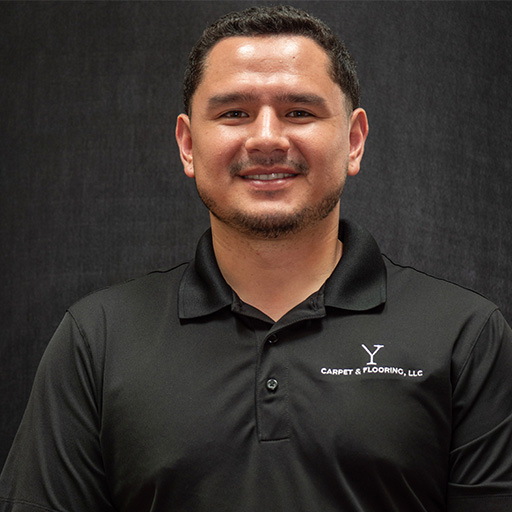 Jorge with short dark hair, trimmed beard, and a friendly smile wears a black collared shirt with a "Carpet & Flooring, LLC" logo on the chest, standing against a dark background.