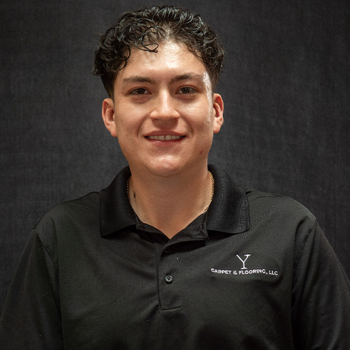 Juan with short, curly dark hair smiles at the camera. He is wearing a black collared shirt with "V Carpet & Flooring, LLC" embroidered on the chest, standing in front of a plain dark background.